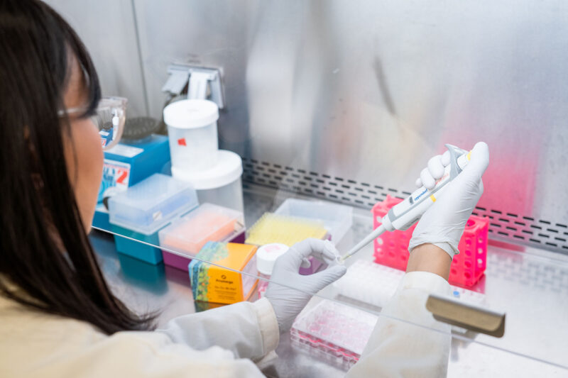 Heather Baker works on her research in the lab, holding a sample and pipette under a fume hood.