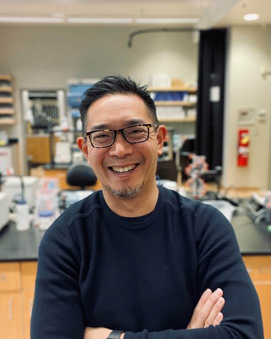 Dr. David Ng, pictured with his arms casually crossed and smiling in front of a lab bench.