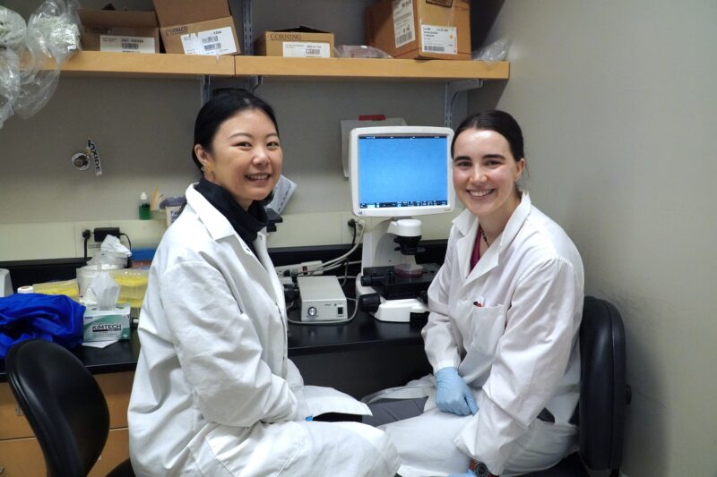 Graduate students Stephanie Besoiu and Clara Xia smile while sitting in front of a microscope in their lab.