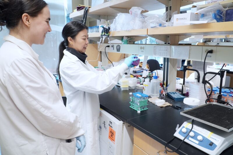 Graduate student Clara Xia pours a sample at a lab bench, joined by Stephanie Besoiu who observes from her side.