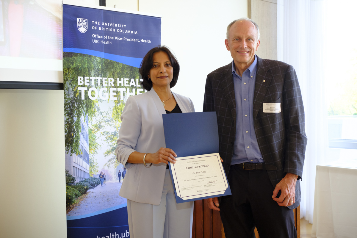 Dr. Brett Finlay stands, smiling, next to Faculty of Pharmaceutical Sciences Dean Dr. Lalitha Raman-Wilms. Dr. Raman-Wilms presents a certificate of award to Dr. Finlay. In the background, a banner reads ‘Better Health Together’ from the Office of the Vice-President for Health at UBC.