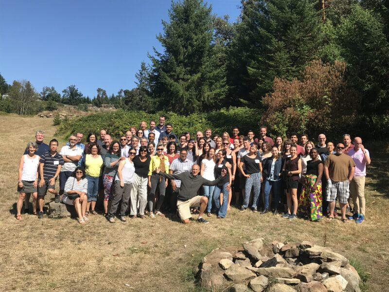 A large group of former Finlay lab members stand together outdoors, smiling to camera. In the very front, Dr. Brett Finlay kneels with his arms out in celebration.